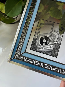A close-up of a stained glass panel with an illustration of a record player sitting in the corner of a room painted in black and gray on a clear piece of glass. Surrounding it is a thin light blue border, then a border of small squares alternating between vertical and horizon black stripes, and a final thin border of darker blue. The while piece is framed in silver zinc.