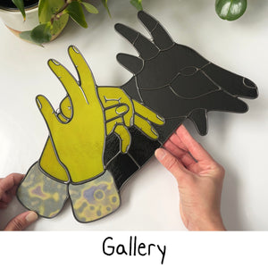 A stained glass depiction of two citronelle green hands with yellow, lavender and gray mottled sleeves forming the shape of a shadow puppet goat, which is displayed to the right in opaque black glass. The word "Gallery" is at the bottom of the image