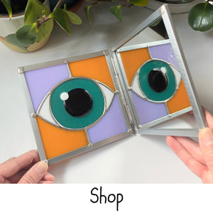 A hinged stained glass design of an eyeball is held open to the camera-- on the left is a fused teal eye surrounded by alternating pumpkin and neolavender background glass. The image of the eye is being reflected by a mirror on the right side. The word "Shop" is at the bottom center of the image