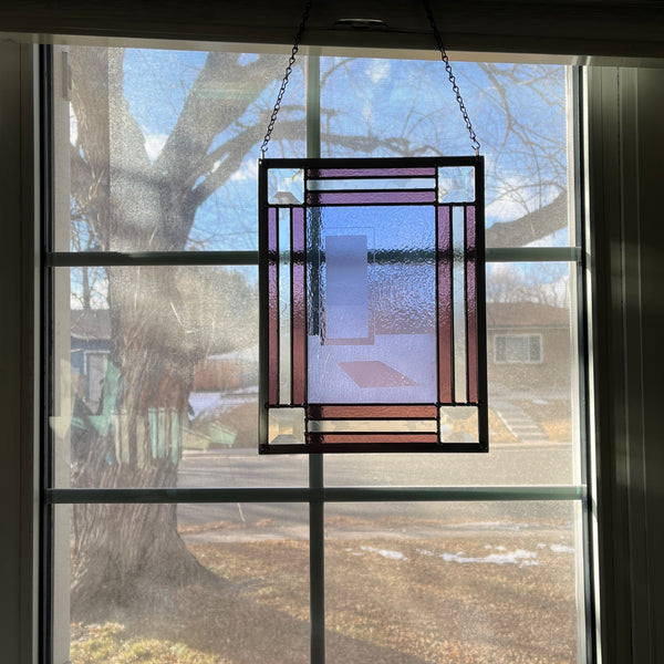 Rectangular stained glass panel hanging in a sunny window depicting a delicate sandblasted graphic of light shining into a glass door, surrounded by clear and deeper purple alternating border with four clear square bevels in each corner