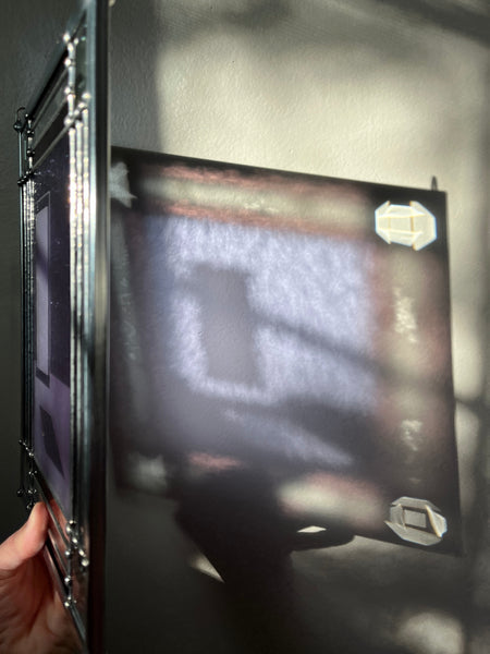 View of the neolavender and darker violet refraction from a rectangular stained glass panel held in front of a khaki colored call with direct light hitting the front. This piece depicts a delicate sandblasted graphic of light shining through a glass door, surrounded by a clear and deeper purple alternating border with four clear square bevels in each corner.